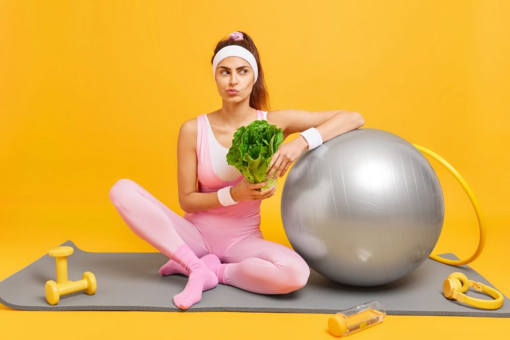 woman-keeps-diet-has-regular-fitness-training-keeping-fit-holds-green-vegetable-dressed-sportsclothes-sits-mat-with-headphones-dumbbells-swiss-ball-hula-hoop-gym